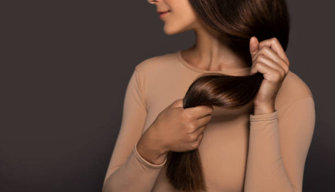 Brunette girl with long straight and shiny hair. Beauty skin woman holding her strong and healthy hair in her hands over grey background. Cosmetic hair beauty salon concept.