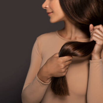 Brunette girl with long straight and shiny hair. Beauty skin woman holding her strong and healthy hair in her hands over grey background. Cosmetic hair beauty salon concept.